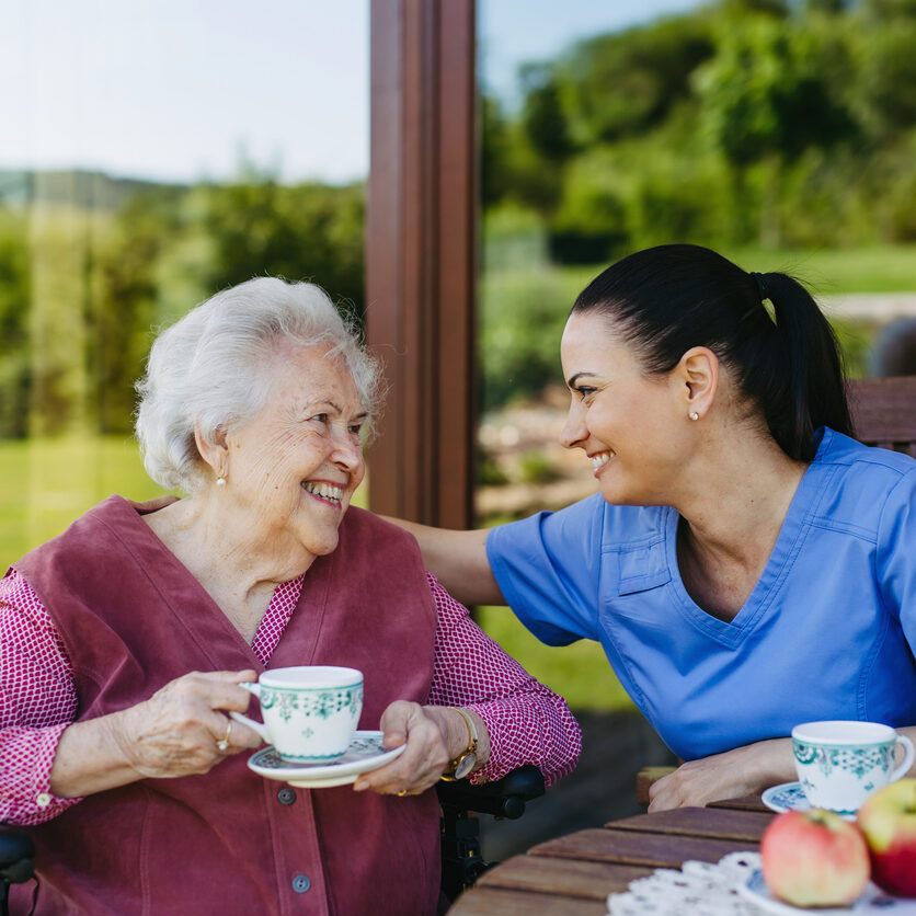 Caregiver spending time with elderly woman, drinking coffee, talking. Nurse and senior woman in wheelchair enjoying a warm day outdoors.